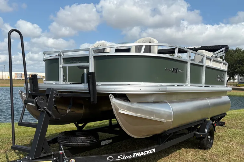 The Image of 2027 Sun Tracker Bass Buggy 16 XL Select pontoon boat on trailer by a lake. - 0