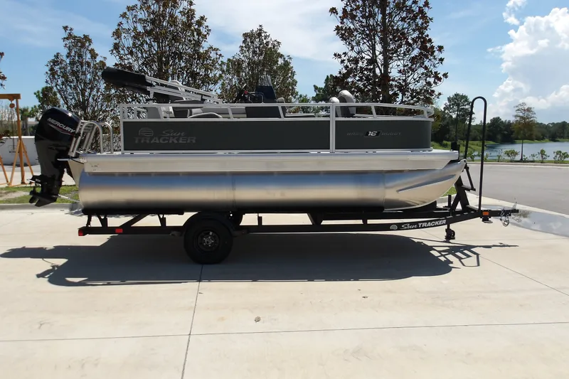 The Image of 2027 Sun Tracker Bass Buggy 16 XL Select pontoon boat on trailer, parked outdoors. - 1