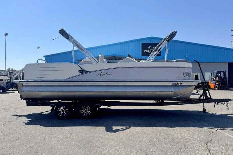 Slide: The Image of 2019 Avalon Catalina Rear Lounge 25' pontoon boat on trailer, parked outside blue building. - 11