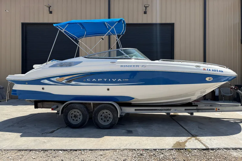 The Image of 2009 Rinker 248 Captiva Deckboat with blue canopy on trailer, parked outside a building. - 0