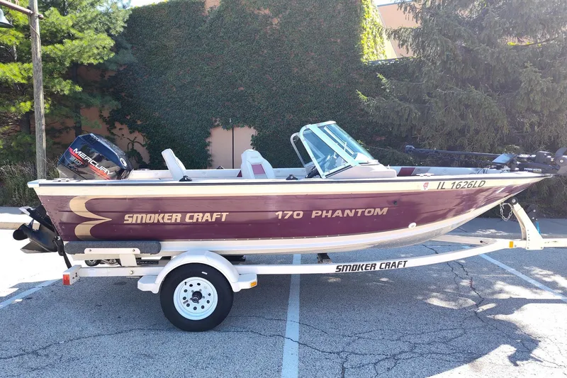 The Image of 1996 Smoker Craft Phantom 16 boat on trailer, parked outdoors with trees in background. - 0