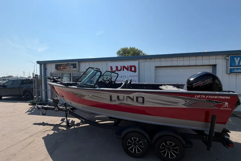 Slide: The Image of 2024 Lund 1875 Fisherman boat on trailer, parked outside dealership under clear blue sky. - 1
