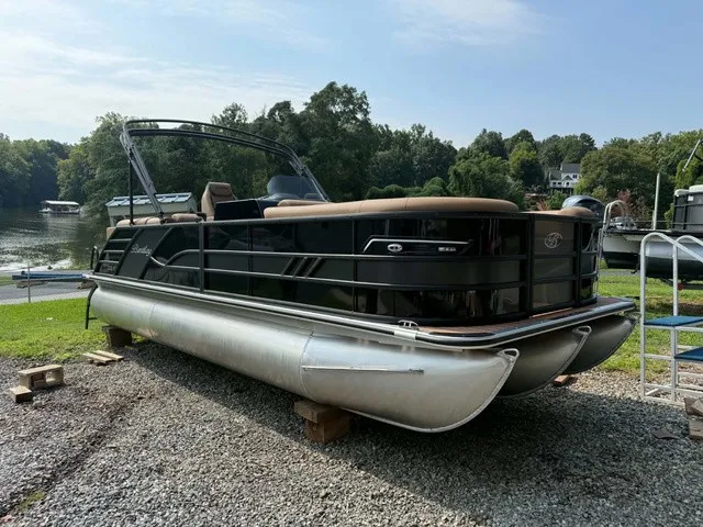 The Image of A sleek pontoon boat with a black hull and beige interior, on a gravel lot by the water. - 1