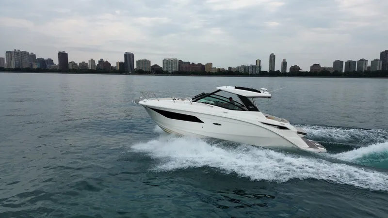 The Image of A 2023 Sea Ray Sundancer 320 speeds across calm waters with a city skyline in the background. - 0