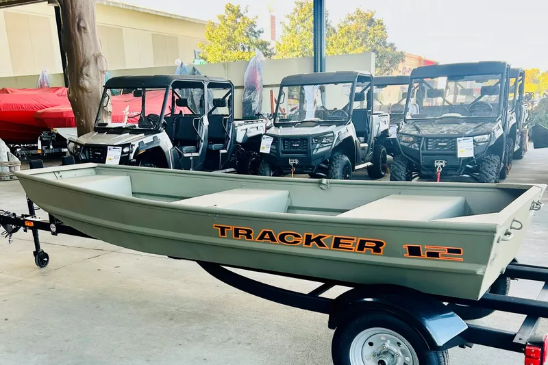 The Image of 2027 Tracker Grizzly 12 Jon boat displayed on trailer, with off-road vehicles in background. - 0