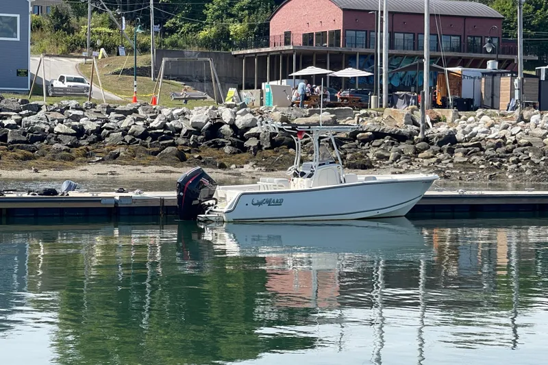 Slide: The Image of 2023 Mako 214 Center Console boat docked at a marina with rocky shoreline. - 23