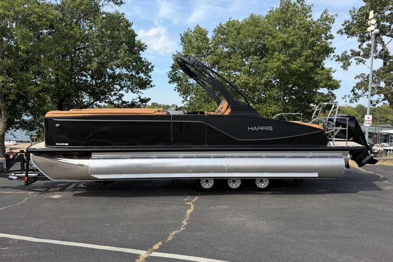 The Image of 2027 Harris Sunliner 250 pontoon boat on trailer, parked near trees and water. - 1
