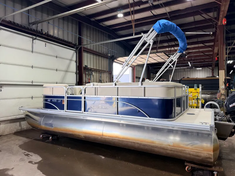 Slide: The Image of A 2015 SunChaser pontoon boat in a warehouse. It features a blue canopy and silver pontoons. - 42