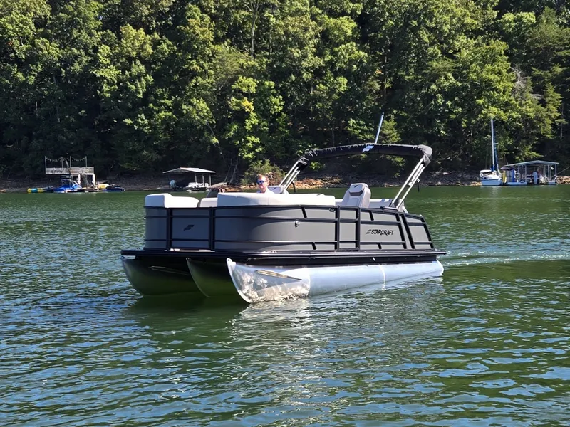 Slide: The Image of A 2027 Starcraft EX 20 Q DH pontoon boat on a calm lake with a wooded shoreline. - 1