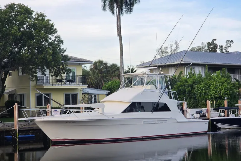 The Image of 1984 Bertram 54 Convertible yacht docked by waterfront homes, surrounded by lush greenery. - 0