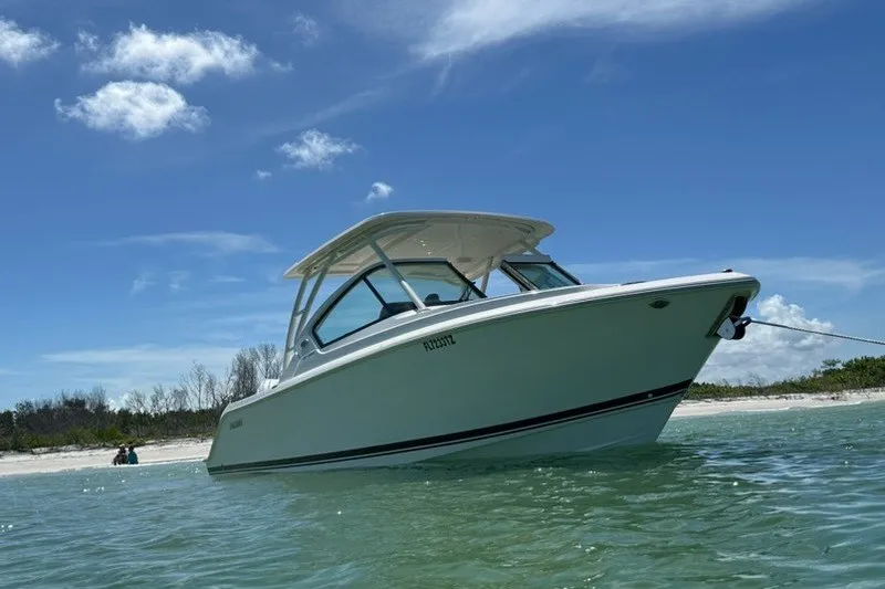 The Image of 2025 Pursuit DC 266 Dual Console boat anchored near a sandy beach under a clear blue sky. - 1