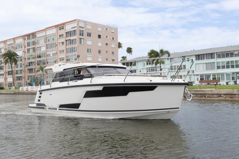 Slide: The Image of 2024 Aquador 300 HT cruising near waterfront buildings on a sunny day. - 2