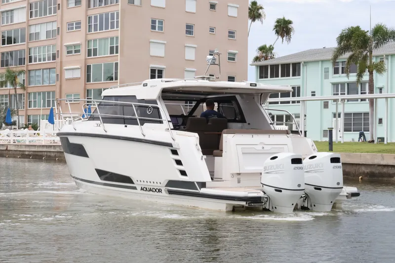 Slide: The Image of 2024 Aquador 300 HT boat cruising near waterfront buildings with twin Suzuki outboard engines. - 13