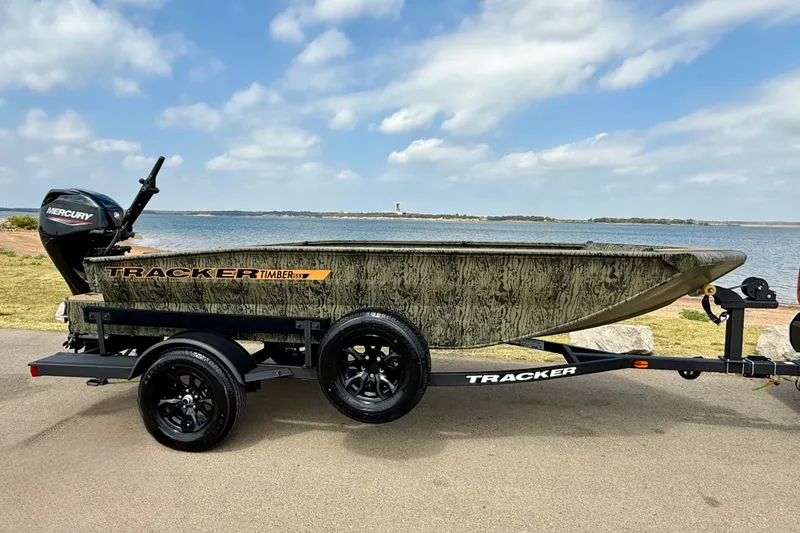 The Image of 2027 Tracker Grizzly 1553 Timber boat on trailer by a lake, under a blue sky. - 0