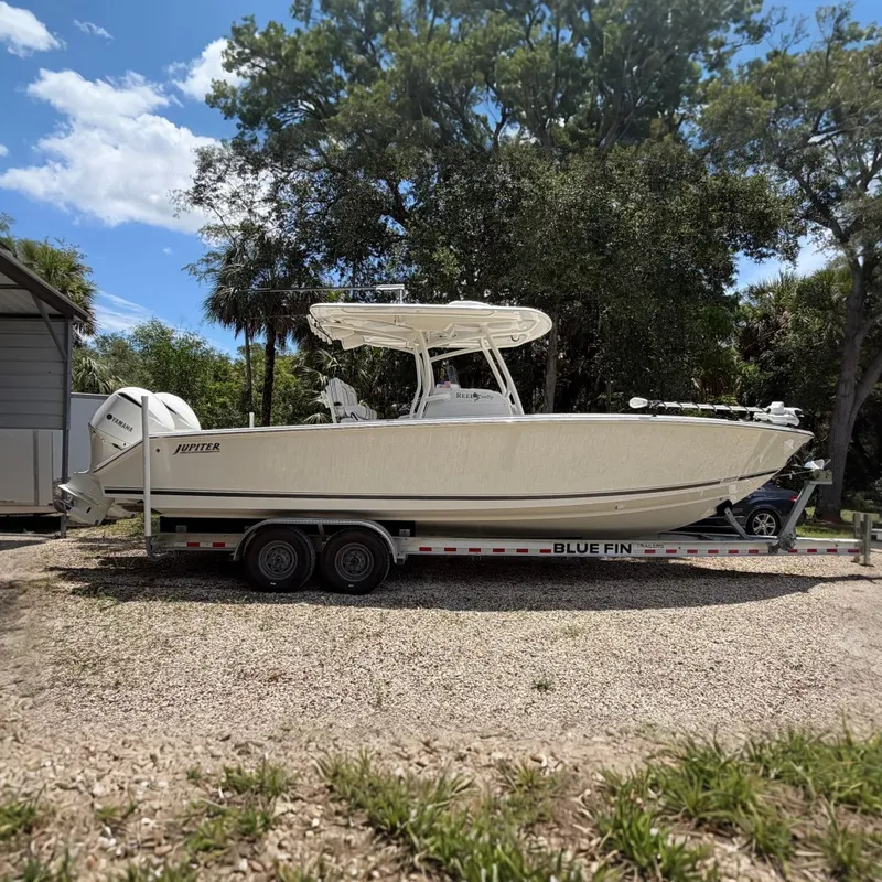 Slide: The Image of 2019 Jupiter 30 boat on Blue Fin trailer, parked outdoors with trees in the background. - 0