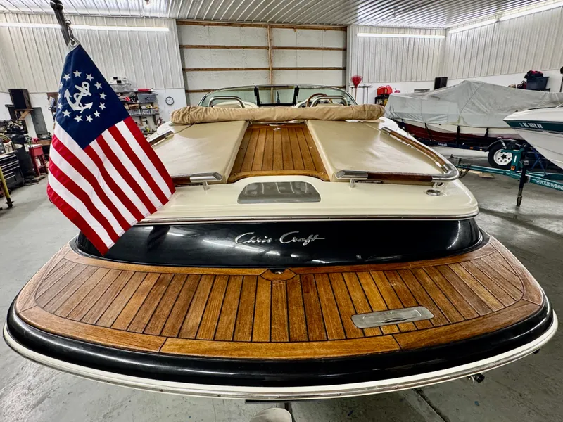 Slide: The Image of A 2016 Chris-Craft Carina 21 boat in a warehouse. It features a sleek wooden deck and an American flag. - 17