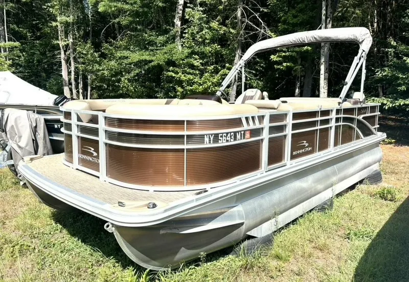 The Image of A 2023 Bennington pontoon boat is on a grassy area, surrounded by trees. - 1