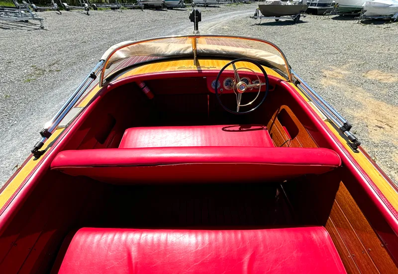 Slide: The Image of A 1956 Chris-Craft Capri with red interior and classic wood design, parked on a gravel lot. - 17