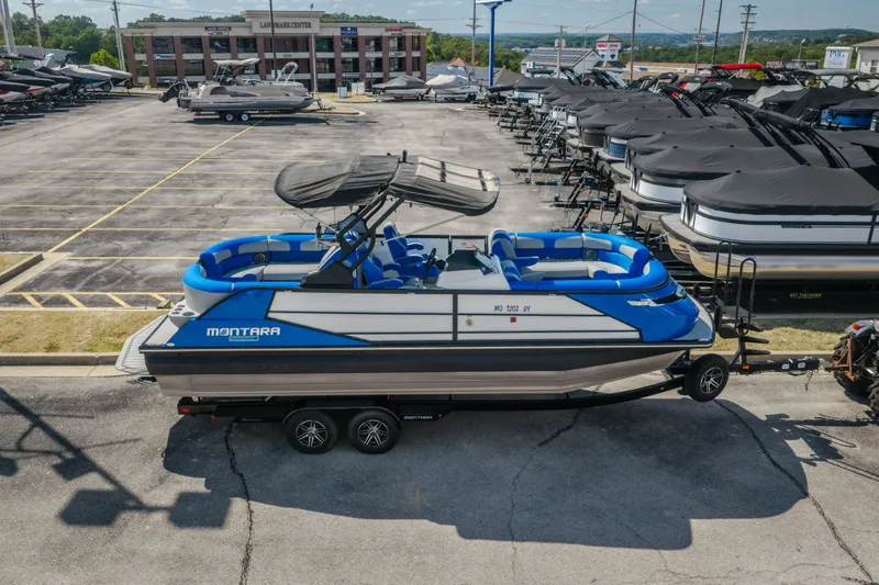 Slide: The Image of 2025 Montara 25 Surf Boss boat in dealership lot, blue and white design, on trailer. - 7