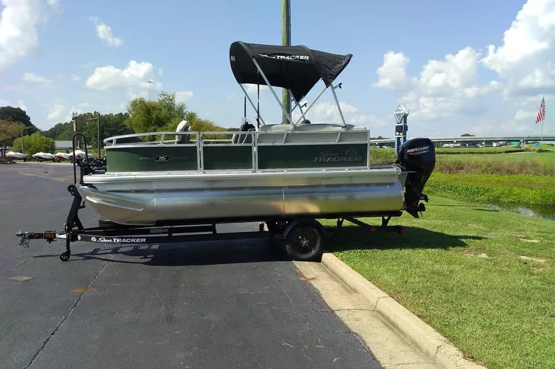 The Image of 2027 Sun Tracker Bass Buggy 16 XL Select pontoon boat on trailer, parked outdoors. - 0