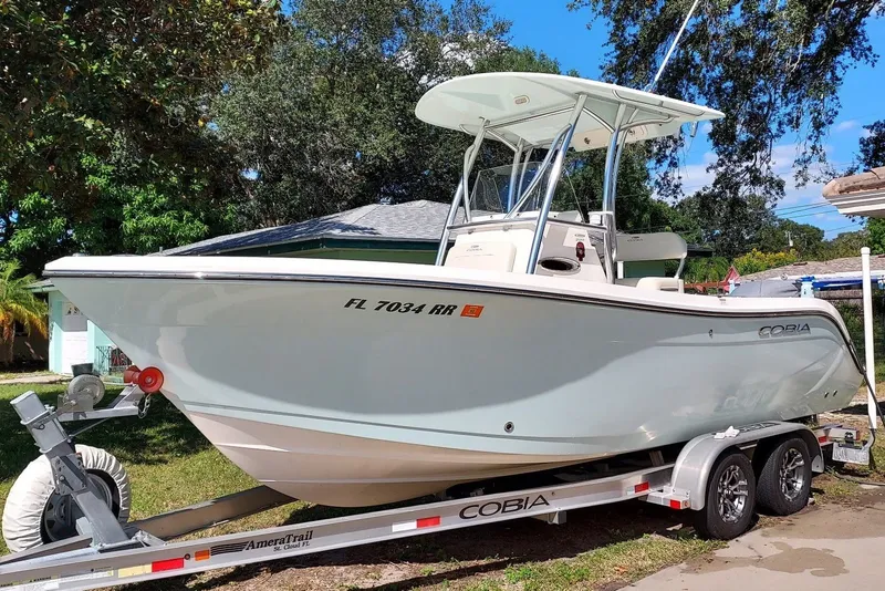The Image of A 2018 Cobia 220 Center Console boat on a trailer, surrounded by trees and a house. - 0