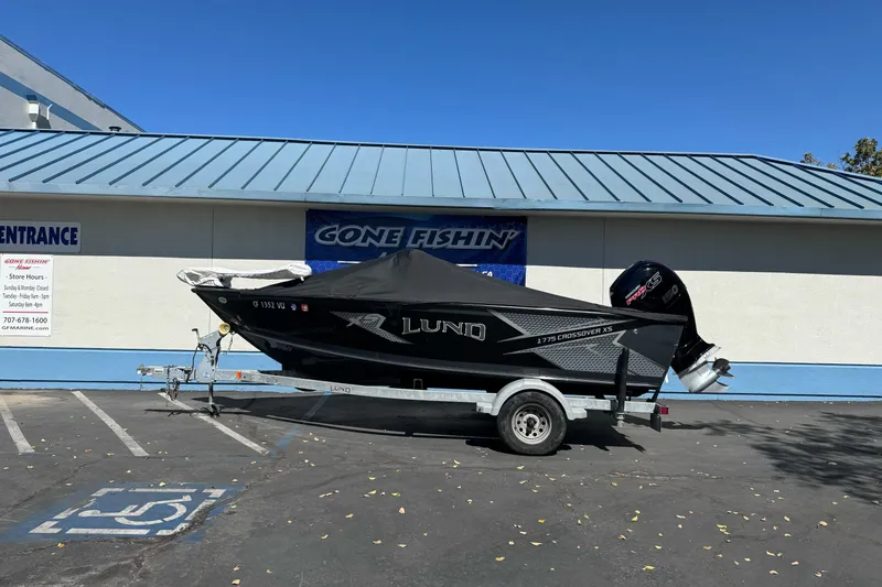 Slide: The Image of 2021 Lund 1775 Crossover XS boat on trailer, parked outside a building under clear blue sky. - 17