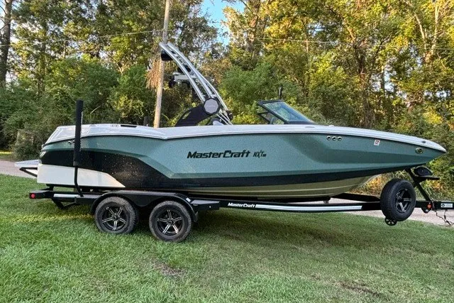 Slide: The Image of 2022 MasterCraft NXT22 boat on trailer, parked on grass with trees in background. - 11