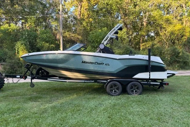 Slide: The Image of 2022 MasterCraft NXT22 boat on trailer, parked on grass with trees in background. - 9