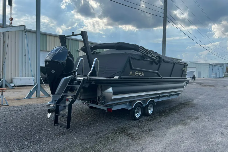 Slide: The Image of 2026 Alera M 23Q boat with cover, parked on a trailer in an industrial area. - 1