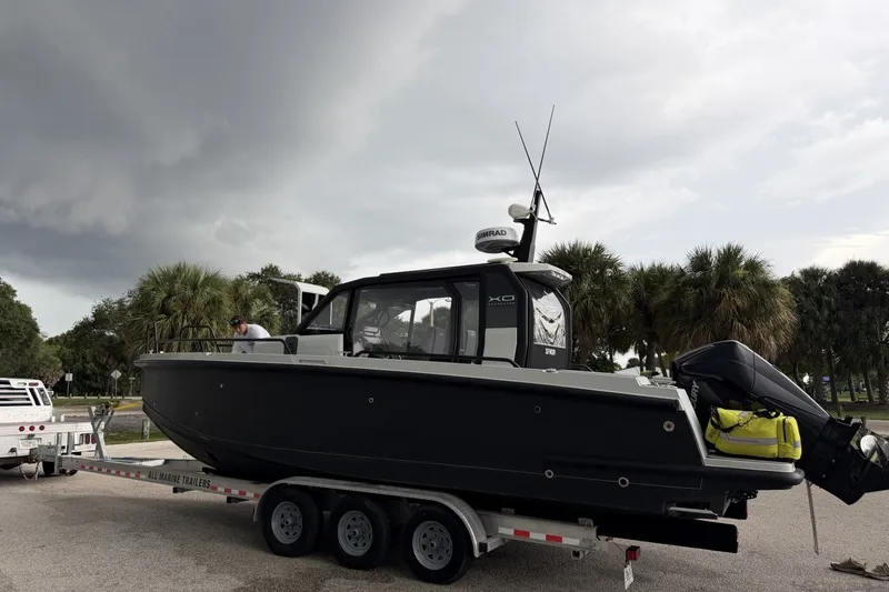 Slide: The Image of 2023 XO DFNDR 9 boat on trailer, parked near palm trees under cloudy sky. - 2