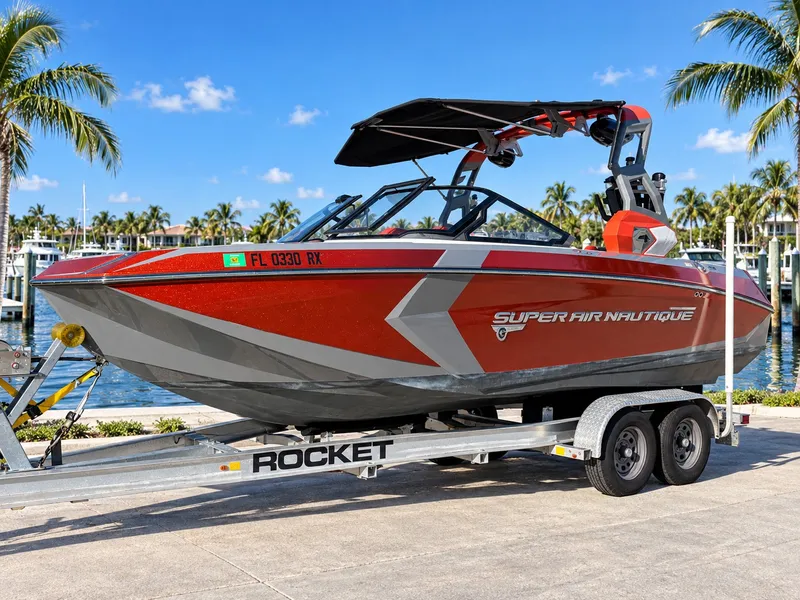 The Image of 2019 Nautique Super Air Nautique G23 on a trailer, surrounded by palm trees and water. - 0