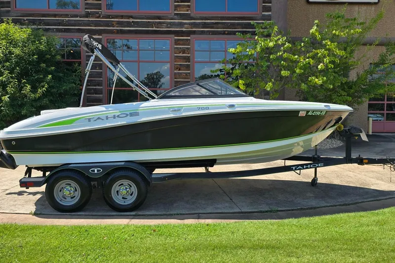 Slide: The Image of 2018 Tahoe 700 boat on trailer, parked outdoors near a building with greenery. - 0