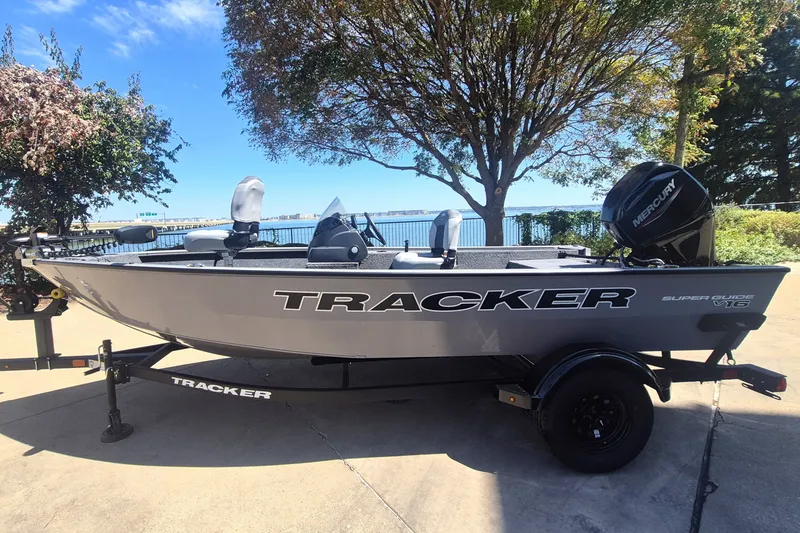 The Image of 2027 Tracker Super Guide V-16 SC boat on trailer, parked outdoors near trees and water. - 0