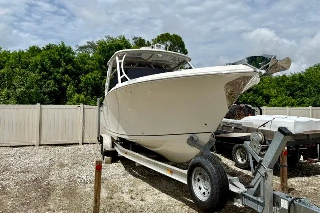 Slide: The Image of 2015 Sailfish 276 DC boat on trailer, parked outdoors with trees and fence background. - 23