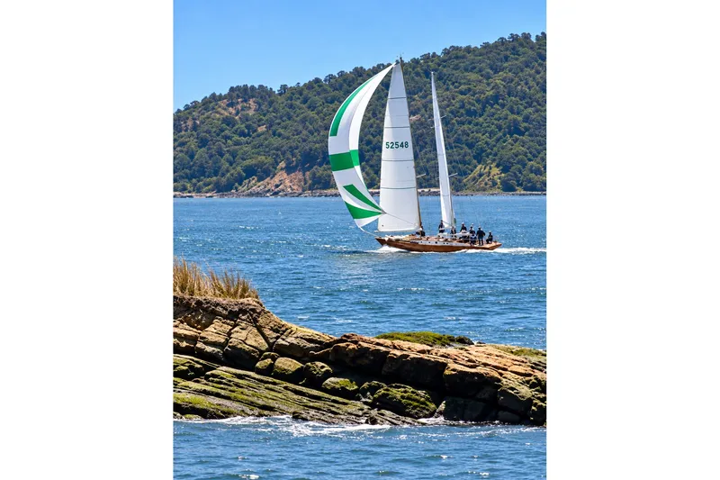 Slide: The Image of Sailing schooner "Chuck Burns Custom 49" gliding on blue waters near rocky shore. - 1
