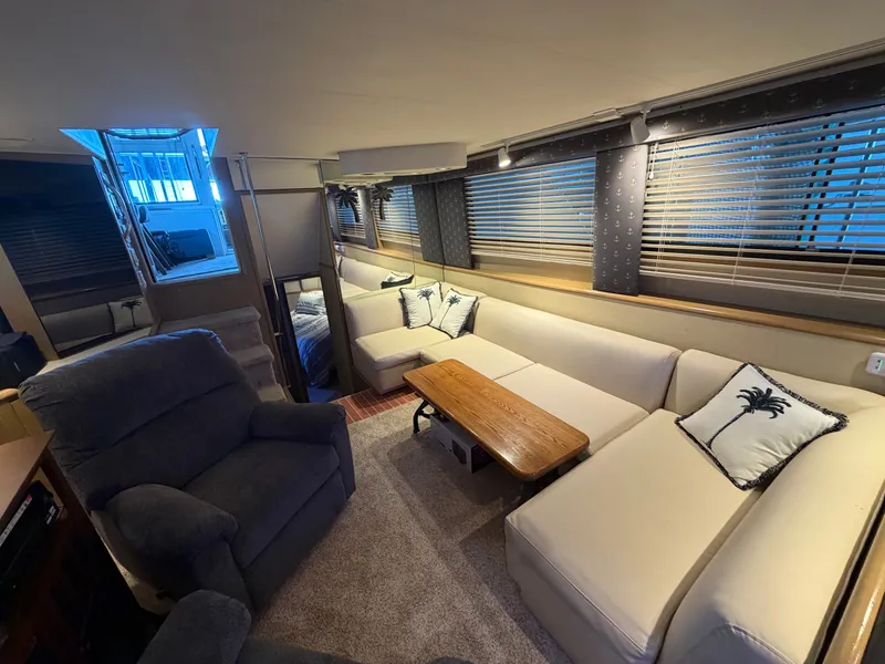 Slide: The Image of The image shows the interior of a 1991 Hatteras 52 Cockpit Motor Yacht. There's a cozy seating area with a large sofa and a wooden table. The windows have blinds, and there are decorative pillows with palm motifs. - 35