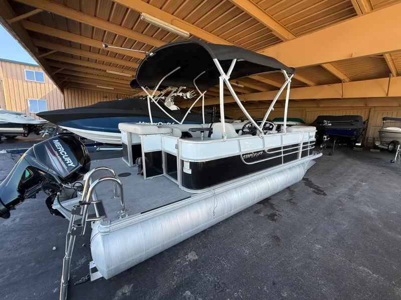 Slide: The Image of A 2017 Starcraft EX 20 C pontoon boat with a black canopy, parked in a covered area. - 2
