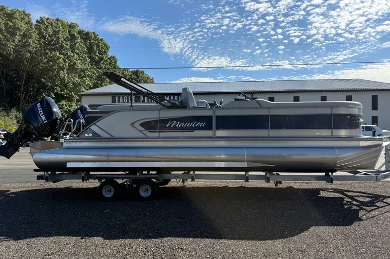 The Image of A Manitou Encore 25 RF Dual pontoon boat on a trailer in front of a building. - 0