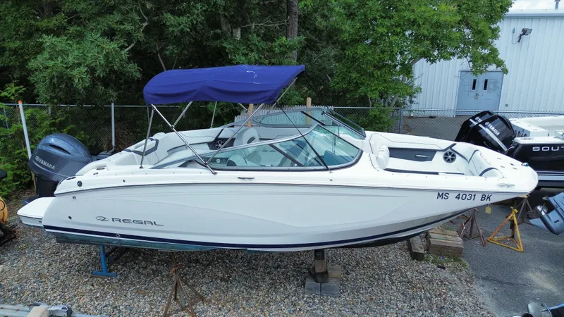 Slide: The Image of A white 2018 Regal 23 OBX boat is on a gravel lot. It features a blue canopy. - 21