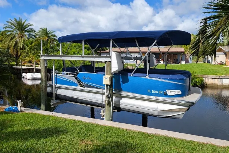 The Image of 2019 Avalon 2485LSZ pontoon boat on lift by canal, surrounded by palm trees. - 0