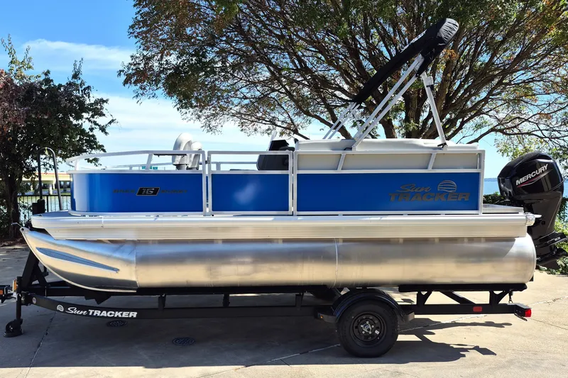 The Image of 2027 Sun Tracker Bass Buggy 16 XL Select pontoon boat on trailer, parked outdoors. - 0