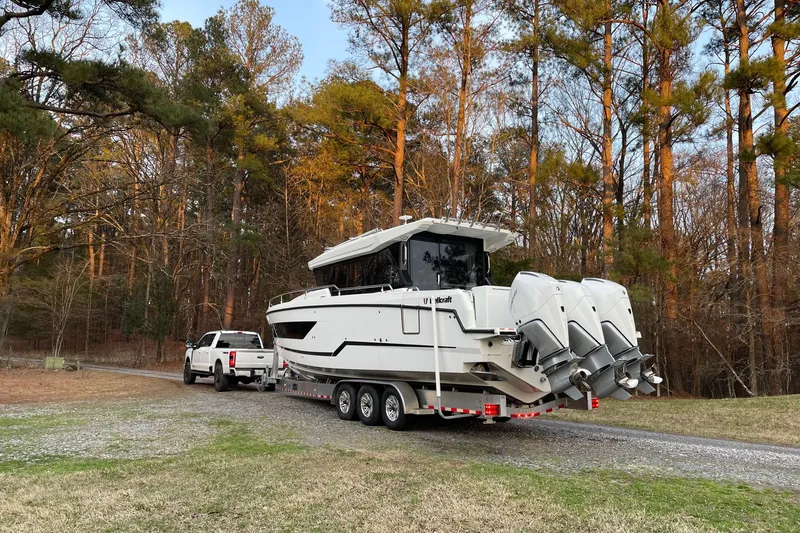 Slide: The Image of 2024 Wellcraft 38 Explorer boat on trailer, towed by a white truck in a wooded area. - 1