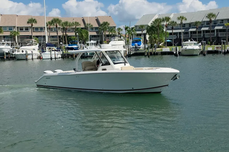 Slide: The Image of 2019 Pursuit S 328 Sport boat cruising in a marina with docked boats and palm trees. - 19