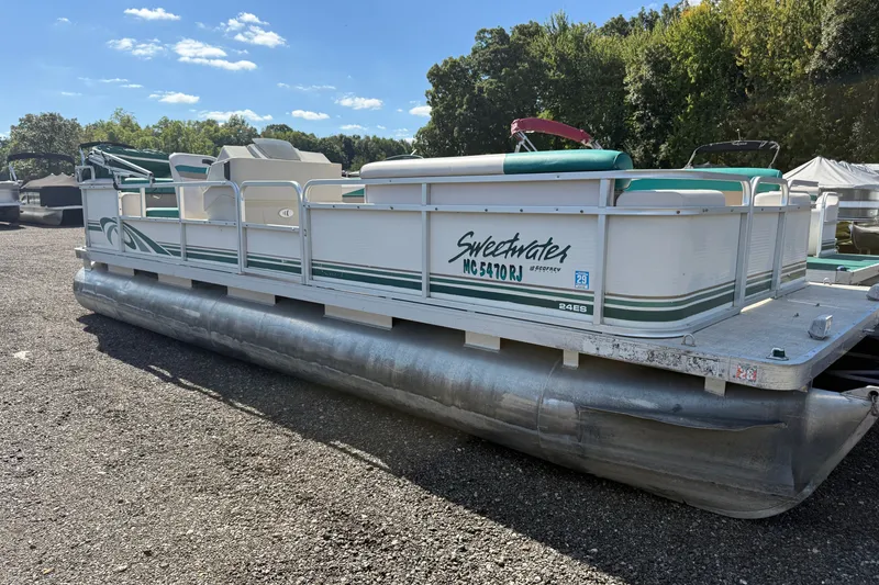 The Image of 1997 Sweetwater 24es pontoon boat parked on gravel, surrounded by trees under a clear sky. - 0