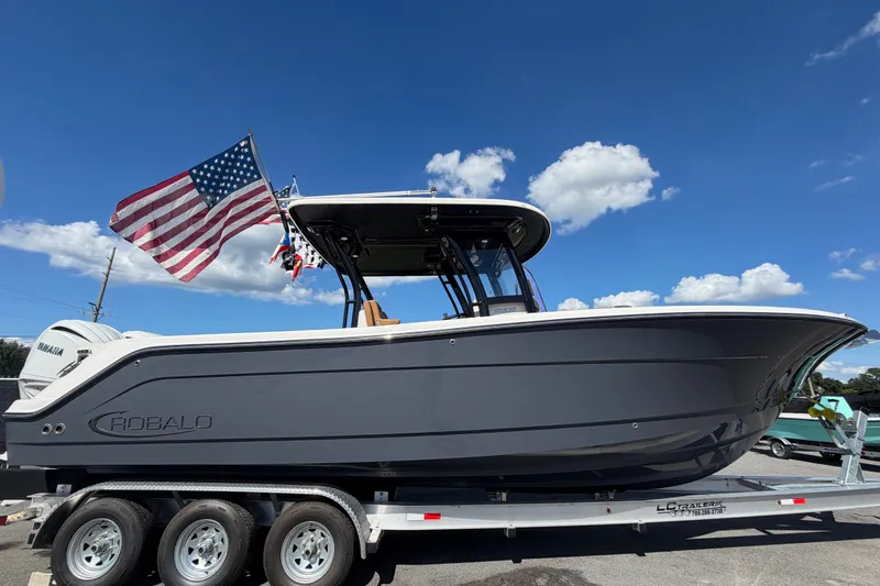 Slide: The Image of 2025 Robalo R302 Center Console boat on trailer with American flag, under clear blue sky. - 2