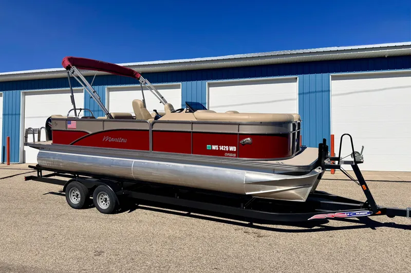 Slide: The Image of 2021 Manitou 23 Oasis pontoon boat on trailer, parked near blue building. - 2