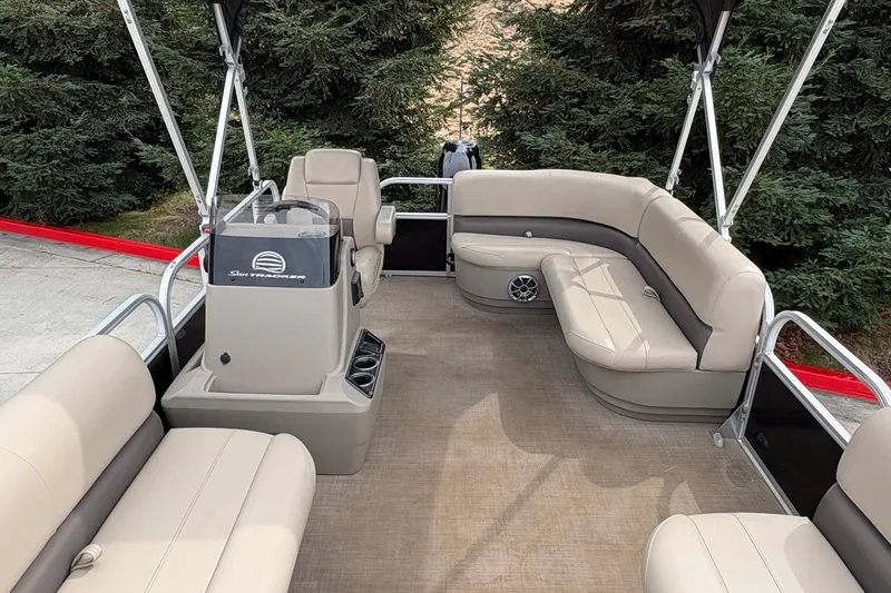 Slide: The Image of 2025 Sun Tracker Party Barge 16 DLX interior with beige seating and helm, surrounded by trees. - 22