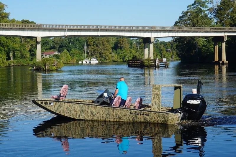 Slide: The Image of Man driving a 2025 Lowe Outlet 20 boat under a bridge on a scenic river. - 6