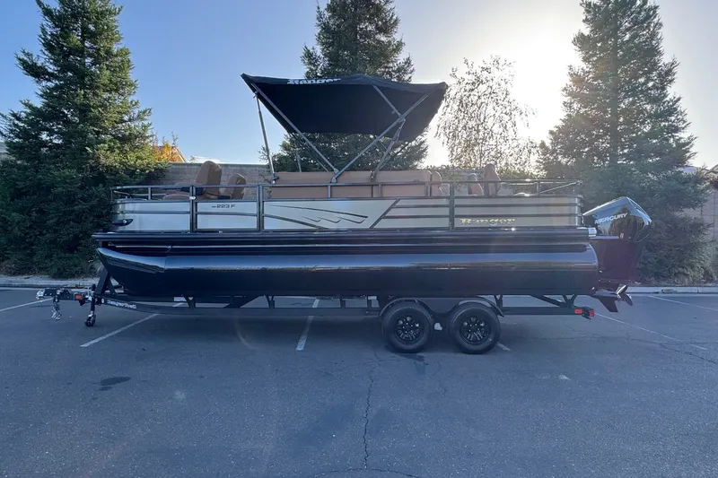Slide: The Image of 2027 Ranger 223F pontoon boat on trailer, parked in a lot with trees in the background. - 1