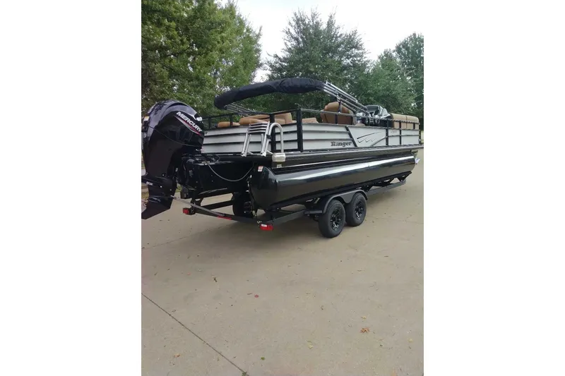 Slide: The Image of 2027 Ranger 220FC pontoon boat on trailer, parked outdoors with trees in background. - 11